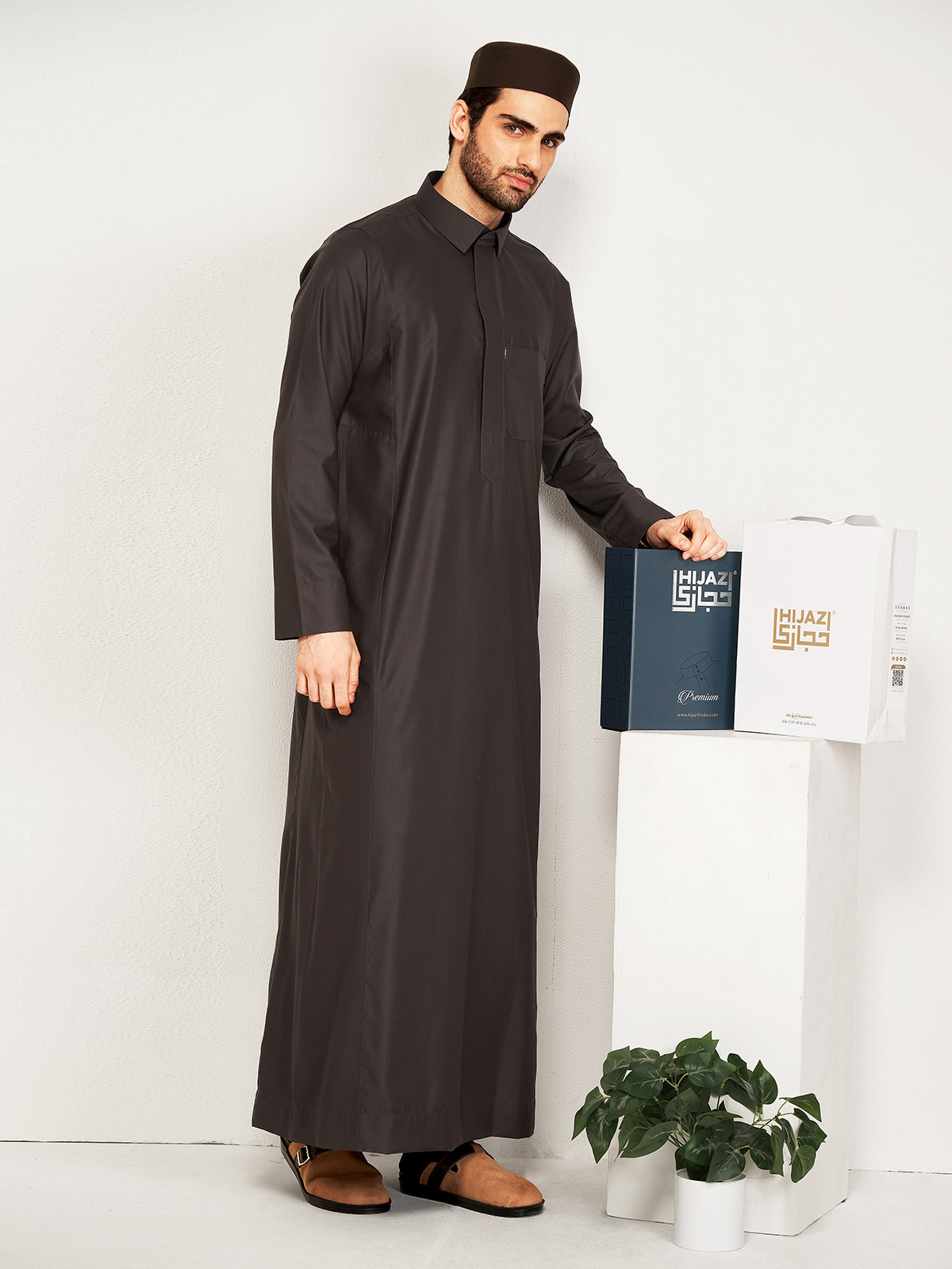 Man wearing a long dark brown robe holding a hijazi product box on a white background