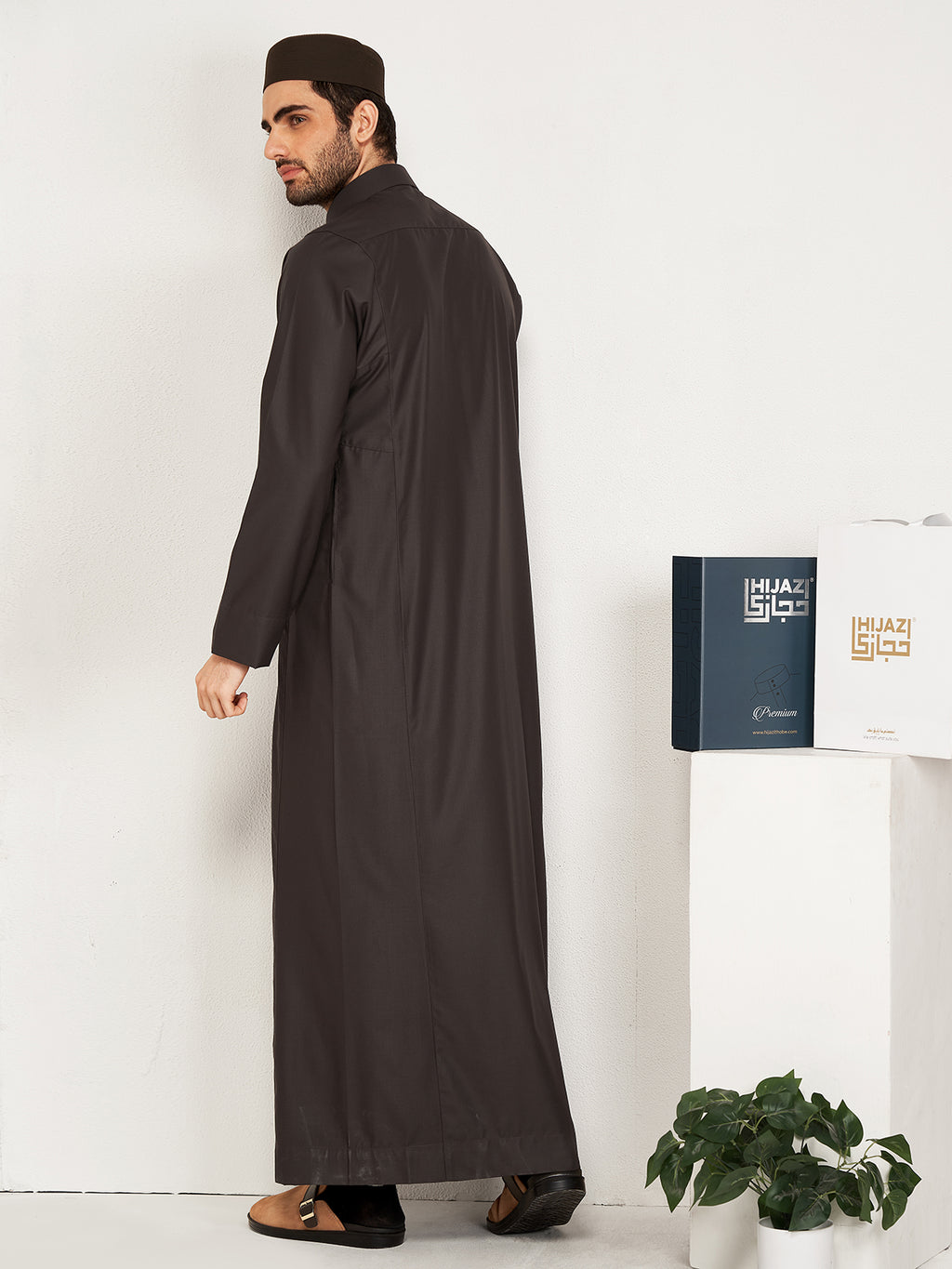 Man wearing a long dark robe standing next to a white wall with a plant and a hijazi box on a table.