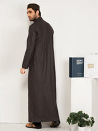 Man wearing a long dark robe standing next to a white wall with a plant and a hijazi box on a table.