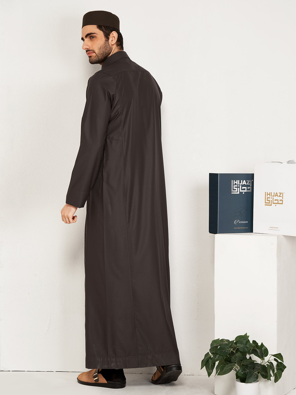 Man wearing a long dark robe standing next to a white wall with a plant and a hijazi box on a table.