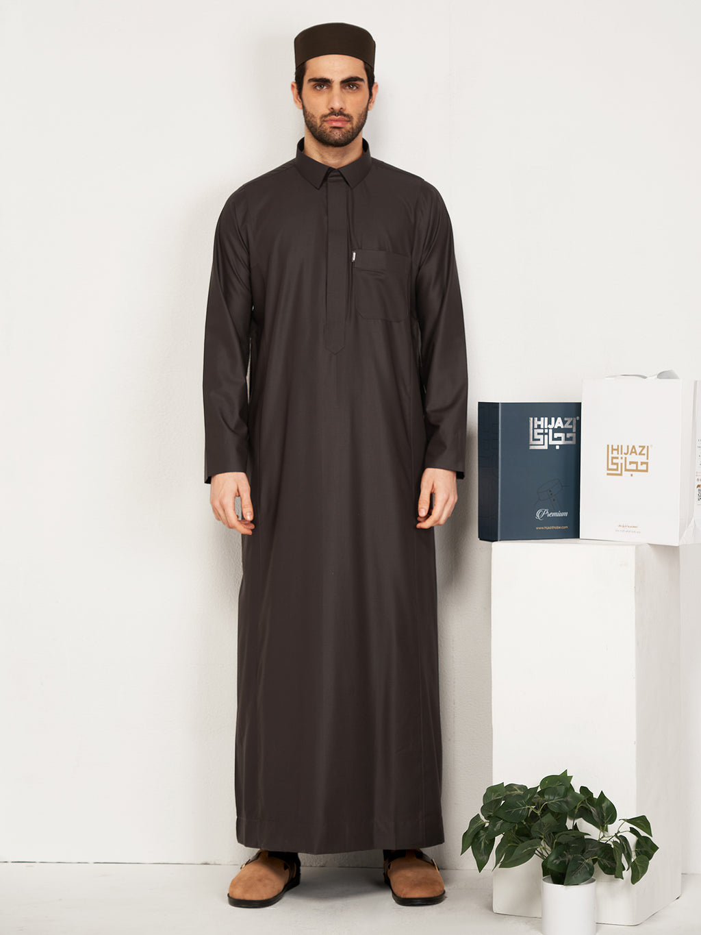  Man wearing a full-length black thobe with traditional cap, holding Hijazi branded boxes against white background