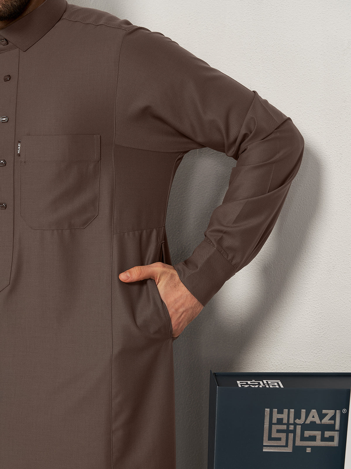 Brown long-sleeve thobeworn by a person against a white background with 'Hijazi' branding.