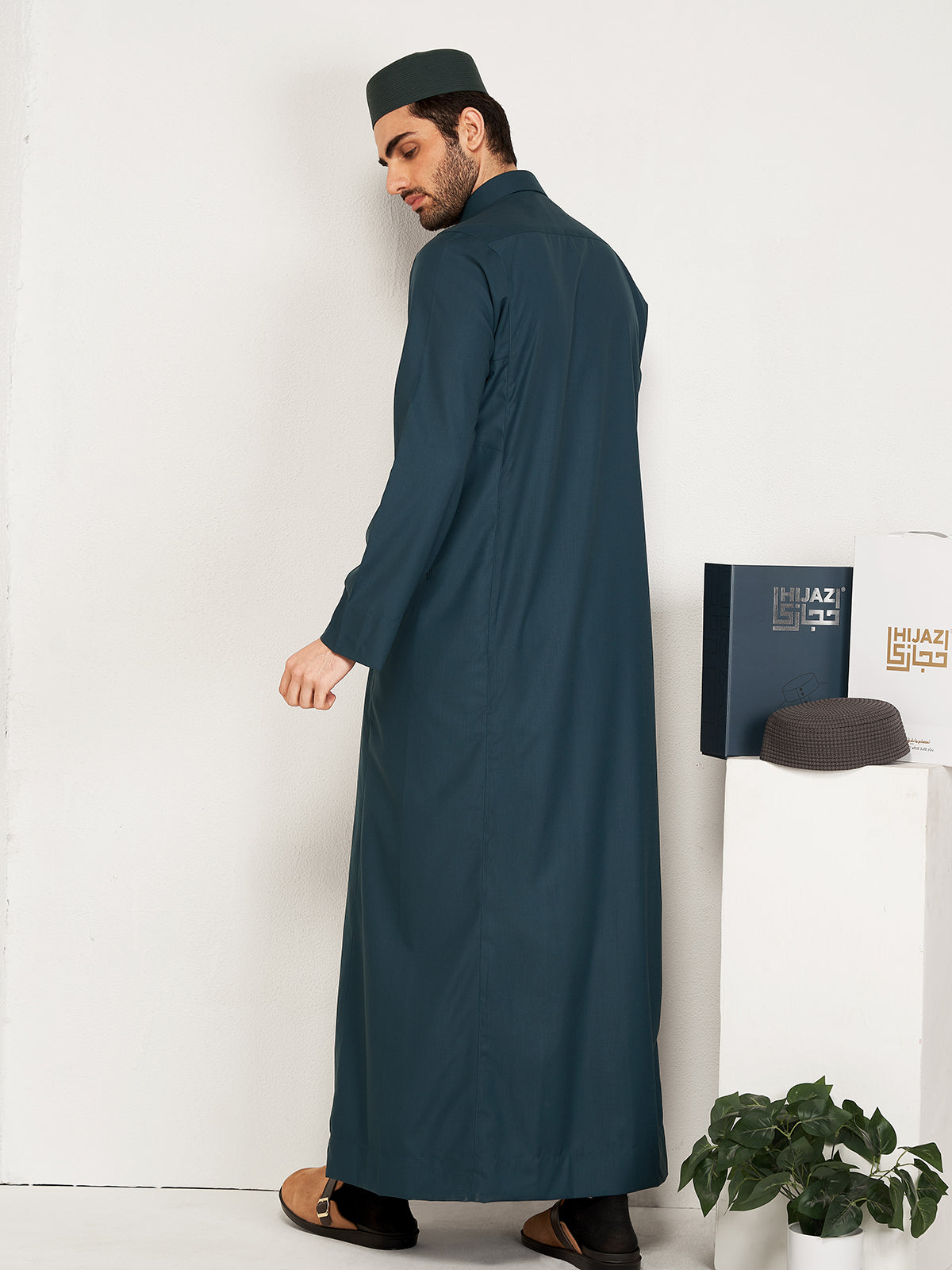 Man wearing a long dark green thobe standing against a white wall with a plant and hijazi box in the background.