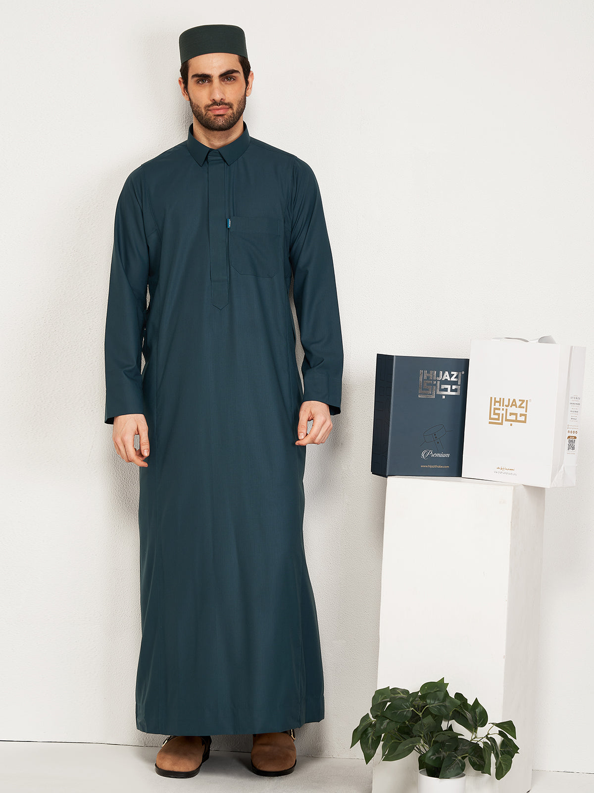 Man wearing a long deep teal thobe standing against a white wall with a plant and a small table in the background.