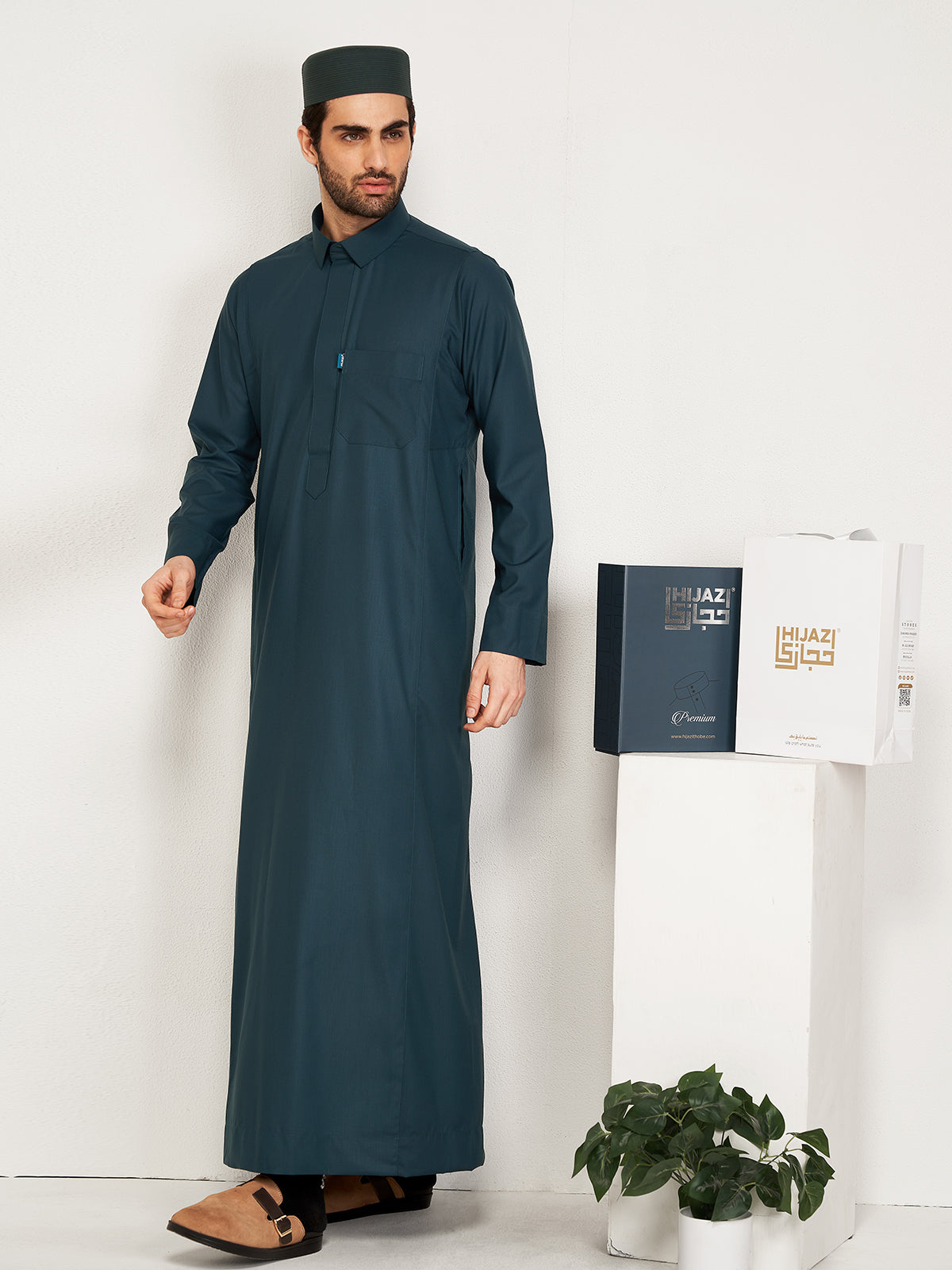 Man wearing a dark green thobe standing next to a white table with a plant and hijazi boxe on it.