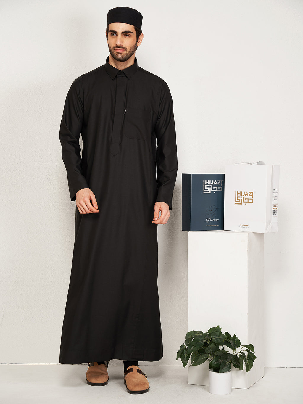 Man wearing a black traditional outfit standing next to a white stand with hijazi box on a white background