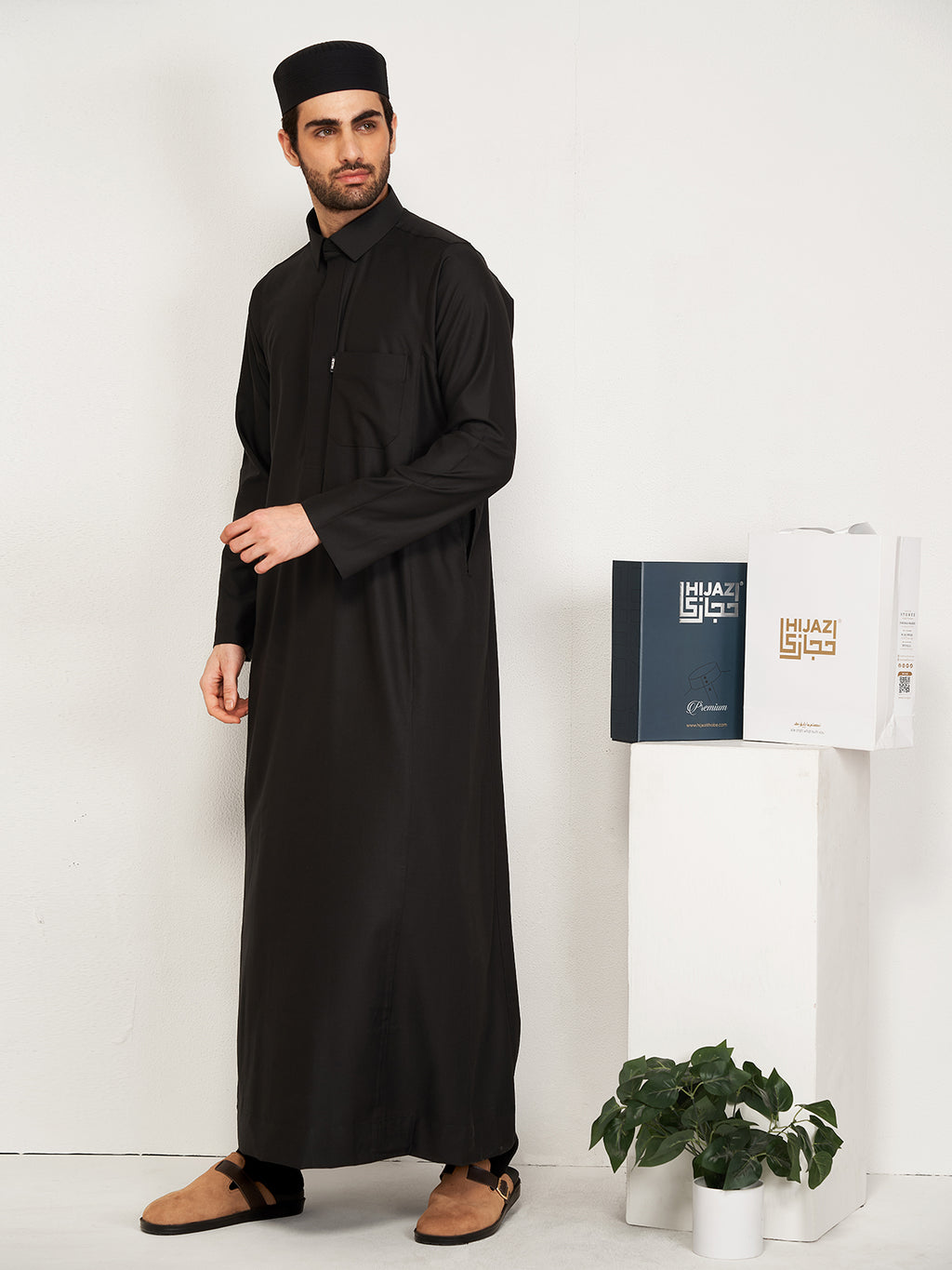 Man wearing a black thobe standing next to a white cabinet with a Hijazi box on a white background