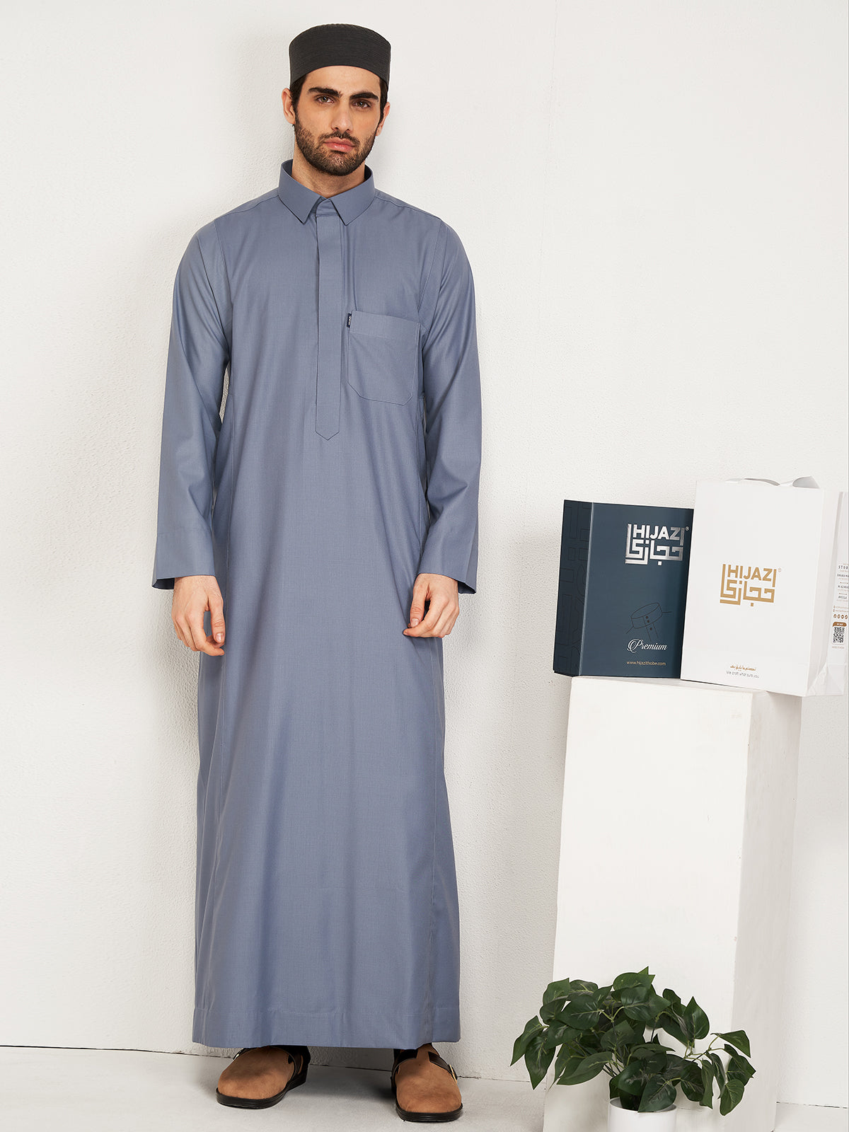 Man wearing a long gray robe standing next to a white wall with a plant and with hijazi box