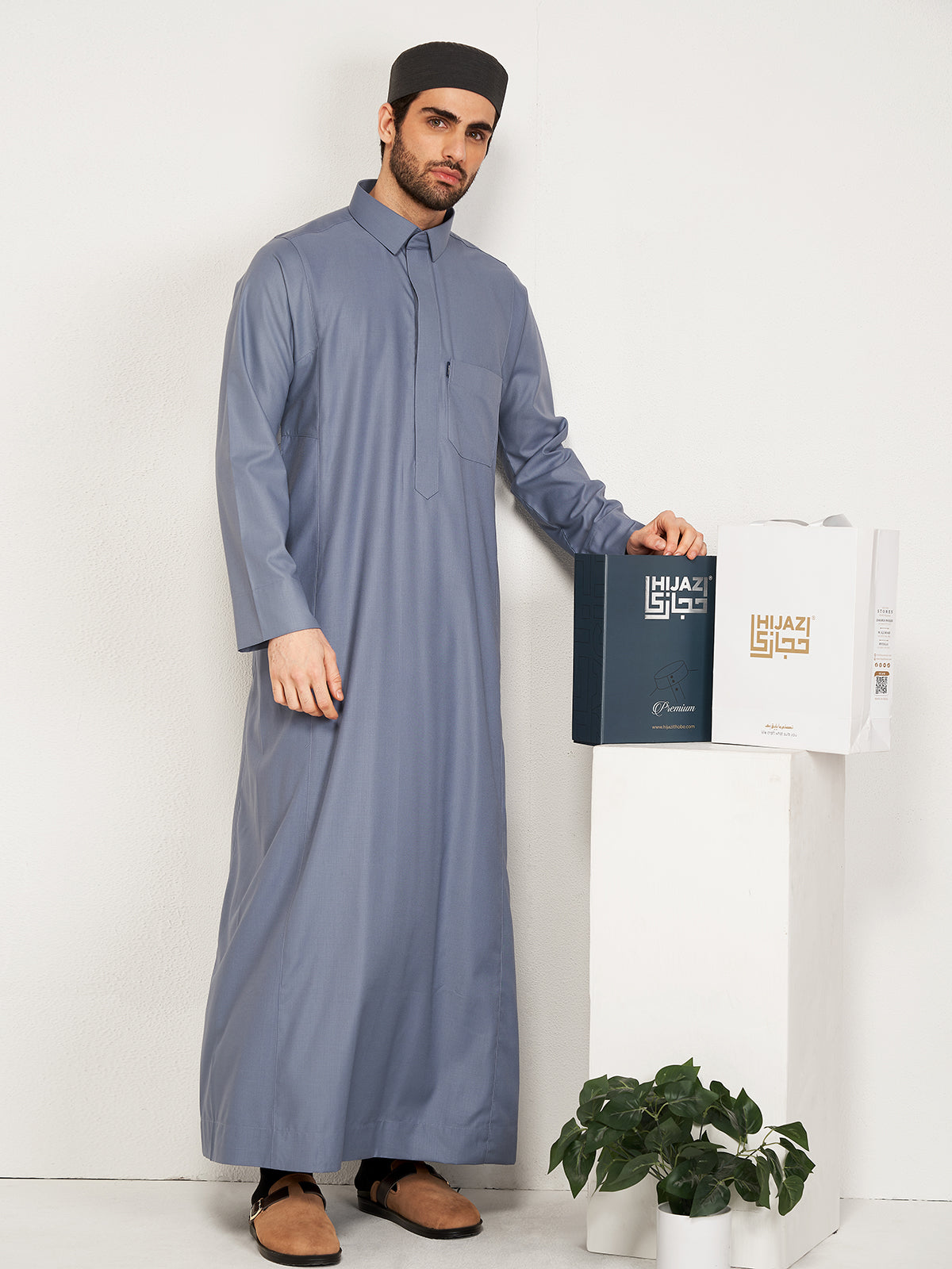 Man wearing a long gray thobe holding a 'Hijazi' product against a white background